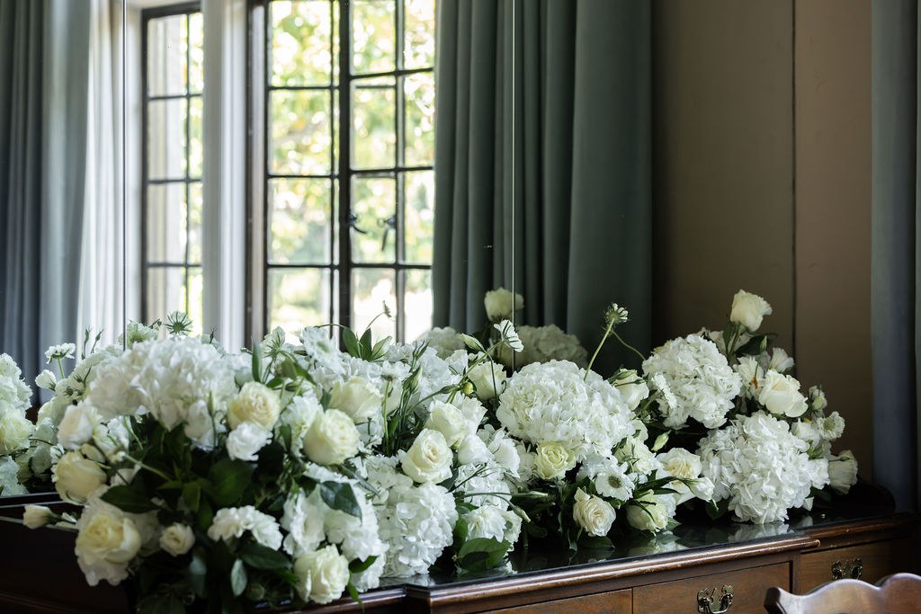 Long white floral arrangement on sideboard by window