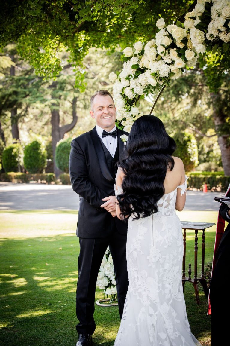 Groom smiling at bride under floral arch on lawn