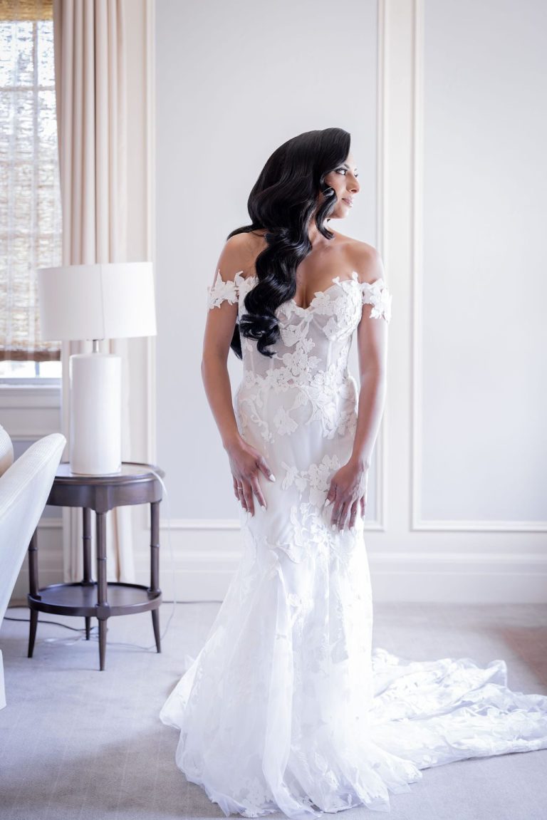 Bride in lace gown standing by lamp table indoors