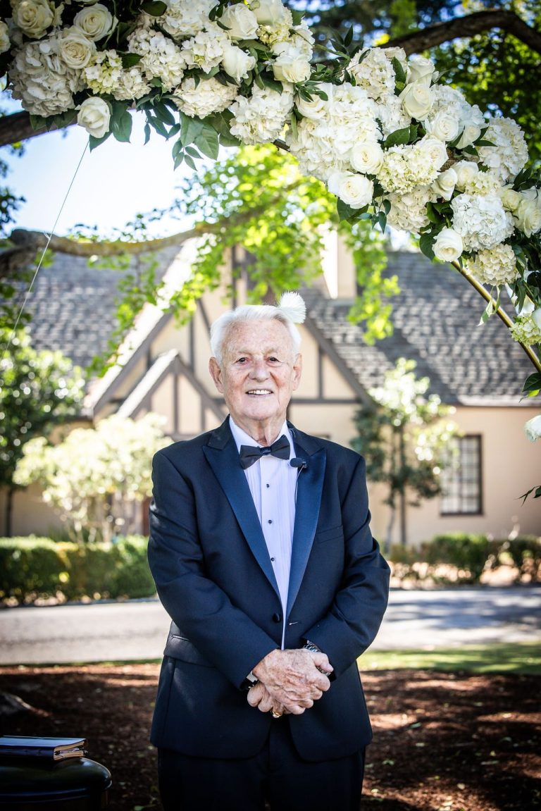 Older man in tuxedo posing beneath floral arch