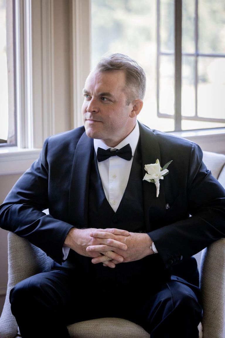 Groom seated leaning forward hands clasped by window