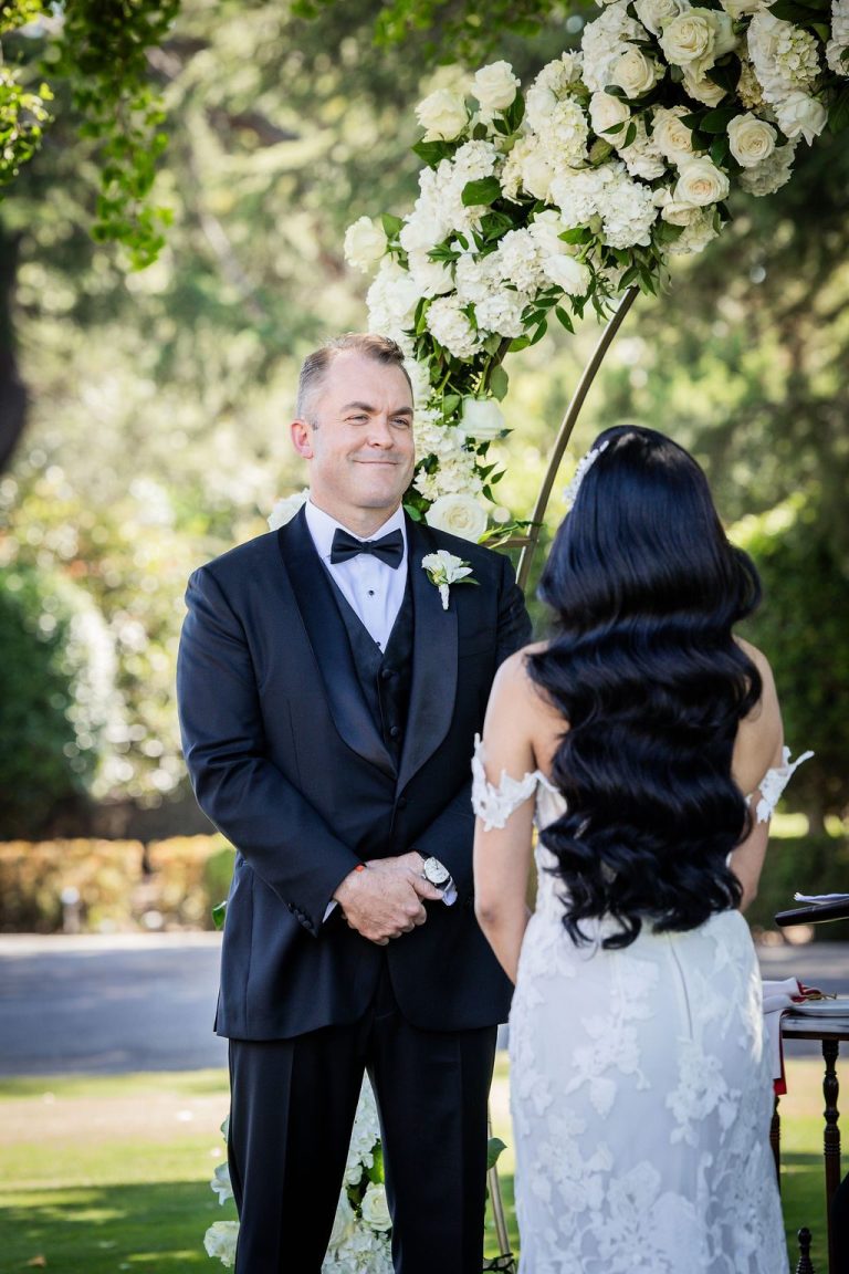 Groom and bride facing each other beneath floral arch
