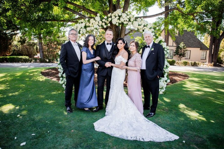 Couple with parents pose at floral arch on lawn