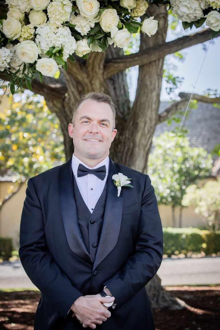 Man in tuxedo smiling beneath white floral arch