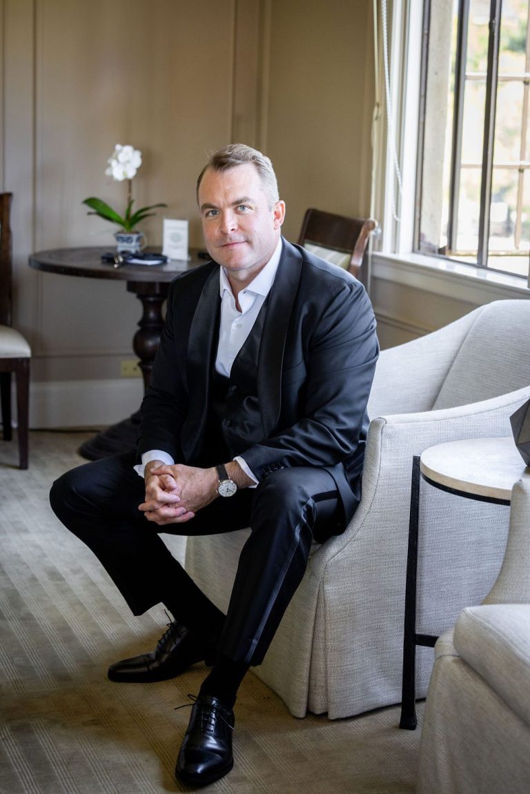 Groom seated smiling with hands clasped by window