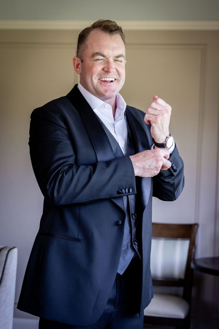 Groom laughing while fixing cufflink in sunlit room