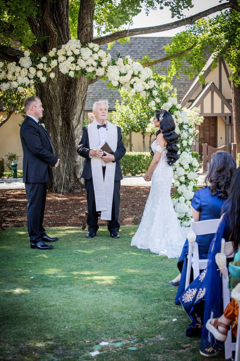 Groom facing bride at arch with guest in foreground