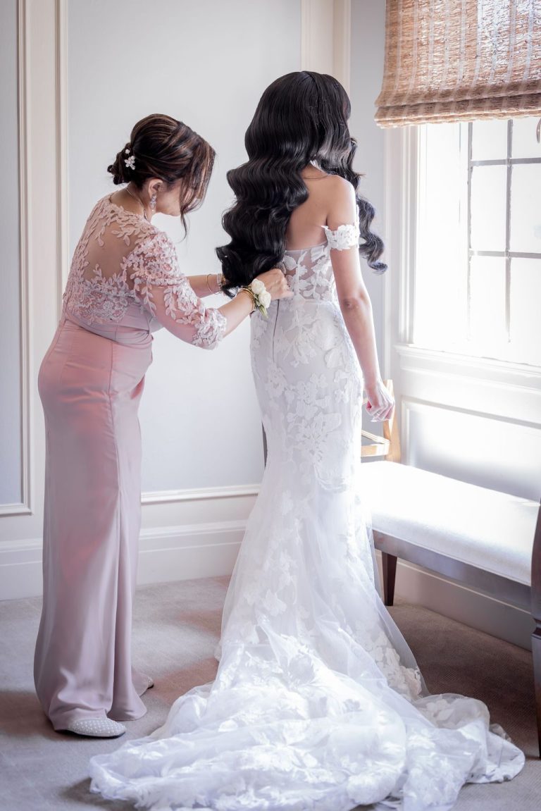 Attendant helping bride with gown near sunlit window