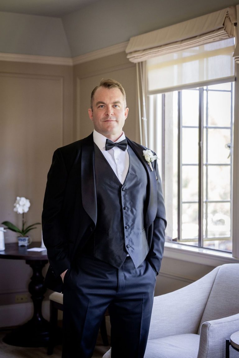 Groom in tuxedo standing confidently by window