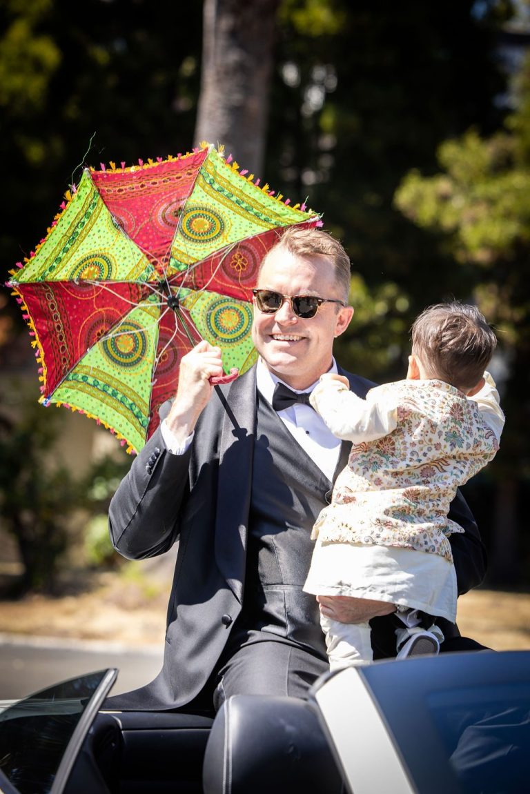 Groom in sunglasses holding child under colorful parasol